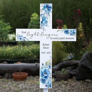 May include: White cross-shaped memorial marker with blue floral accents. The text reads "Your light lingers in every quiet moment." Below, it says "Robert Finch 1946-2025."
