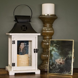 May include: A white wooden lantern with a black top and handle, featuring a glass panel with the text "Keep the Light On" and a photo. Beside it is a gold candle holder with a white candle and a framed painting.