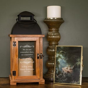 May include: A wooden lantern with a black top and glass window displays a candle and text. Next to it is a tall, ornate candle holder with a white candle and a framed painting with a gold frame. The lantern reads "We do not remember days, we remember moments."