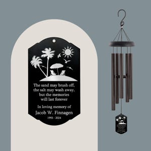 May include: Black metal wind chime with a rectangular plaque featuring a beach scene with palm trees, a beach umbrella, and two lounge chairs. The plaque has the text "The sand may brush off, the salt may wash away, but the memories will last forever. In loving memory of Jacob W. Finnagen 1993-2024."