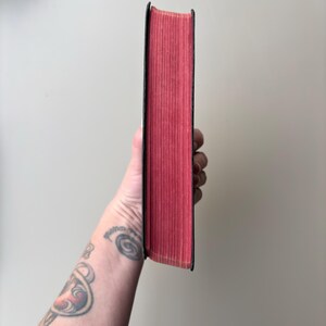 May include: A hardcover book with a black spine and vibrant red page edges. The book is held vertically, showcasing the solid red color of the paper edges against the dark black cover material. The texture of the paper pages is visible.
