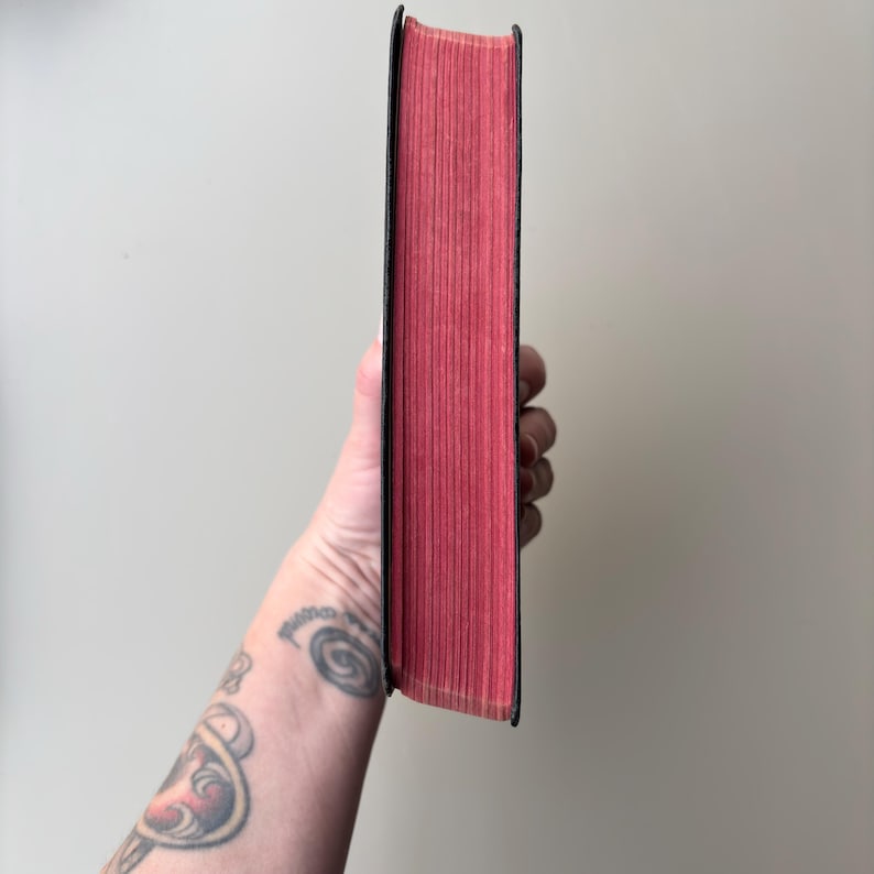 May include: A hardcover book with a black spine and vibrant red page edges. The book is held vertically, showcasing the solid red color of the paper edges against the dark black cover material. The texture of the paper pages is visible.