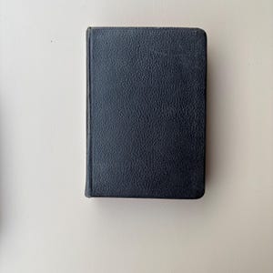 May include: A rectangular book with a dark navy blue cover featuring a textured, pebbled surface. The book is oriented vertically, showing the front cover and the spine edge on the left side. The edges of the cover are rounded at the corners.