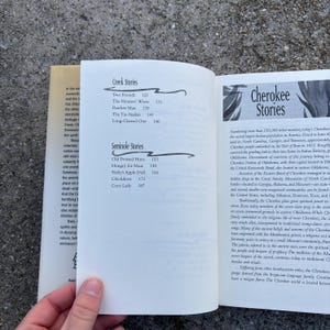 May include: Open book with text about Cherokee and Creek stories. The left page lists "Creek Stories" and "Seminole Stories" with story titles and page numbers. The right page has the title "Cherokee Stories" and detailed text about the Cherokee people.