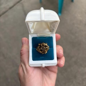 May include: A gold-colored ring with a textured floral design, presented in a white jewelry box with a blue velvet interior. The ring is the focal point, showcasing intricate details.
