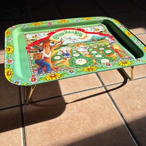 May include: Green metal tray with a Cabbage Patch Kids design. The tray features a colorful illustration of the Cabbage Patch Kids in a garden setting, with the logo at the top. The tray has a floral border and gold-colored legs.