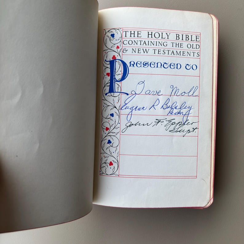 May include: An open Holy Bible with a presentation page featuring red and blue ink text. The page includes the printed words THE HOLY BIBLE CONTAINING THE OLD & NEW TESTAMENTS and PRESENTED TO, followed by handwritten names in blue and black ink.