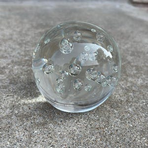 May include: A clear glass sphere with numerous air bubbles suspended within. The spherical object is resting on a textured gray concrete surface, with a brick wall in the background. The glass ball reflects the surrounding environment.