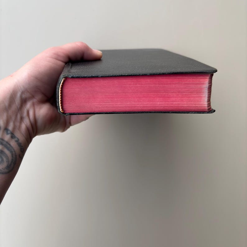 May include: A hardcover book with a textured black cover and vibrant pink page edges. The book is held horizontally, showcasing the contrast between the dark exterior and the bright, solid-colored paper edges that define the thickness of the volume.