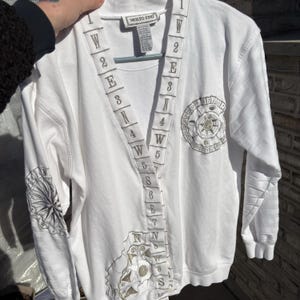 May include: White long-sleeve sweatshirt with embroidered compass rose and directional symbols. The sweatshirt features a V-neck design with embroidered numbers and letters. The brand tag reads "MOLTO FINO".