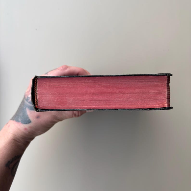 May include: A hardcover book viewed from the side, showcasing a black spine and cover edges with vibrant, solid red-dyed page edges. The book is held horizontally, displaying the uniform, smooth texture of the red paper block against the dark binding.