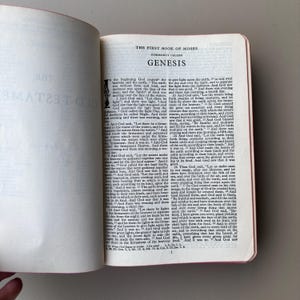 May include: An open book displaying the text of The First Book of Moses, commonly called Genesis. The page features black printed text on a white background with a decorative initial letter at the start of the chapter and numbered verses.