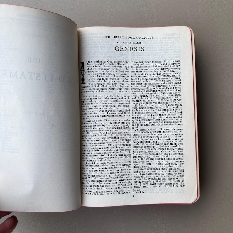 May include: An open book displaying the text of The First Book of Moses, commonly called Genesis. The page features black printed text on a white background with a decorative initial letter at the start of the chapter and numbered verses.