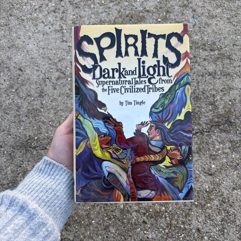 May include: A hardcover book titled "Spirits Dark and Light" with the subtitle "Supernatural Tales from the Five Civilized Tribes" by Tim Tingle. The cover features a colorful illustration of figures in a tribal setting, with a yellow background and bold lettering.