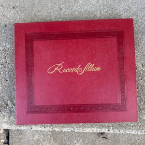 May include: A red record album with gold lettering that reads "Record Album". The album has a decorative border around the edges. The album is rectangular and appears to be made of a leather-like material.