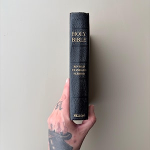 May include: A black, textured hardcover Holy Bible with gold lettering on the spine. The text reads HOLY BIBLE, REVISED STANDARD VERSION, and NELSON. The book is held vertically, showcasing the spine with its distinct horizontal embossed lines.