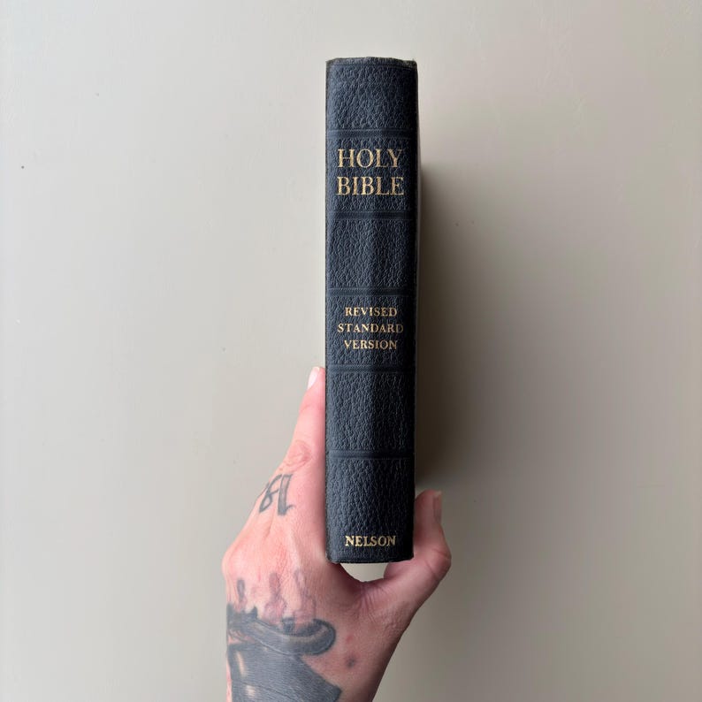 May include: A black, textured hardcover Holy Bible with gold lettering on the spine. The text reads HOLY BIBLE, REVISED STANDARD VERSION, and NELSON. The book is held vertically, showcasing the spine with its distinct horizontal embossed lines.