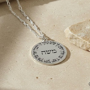 May include: A silver necklace with a circular pendant inscribed with Hebrew script. The pendant is suspended from a fine chain. A small, pale stone sits close by on a neutral surface. The necklace is a piece of jewellery.