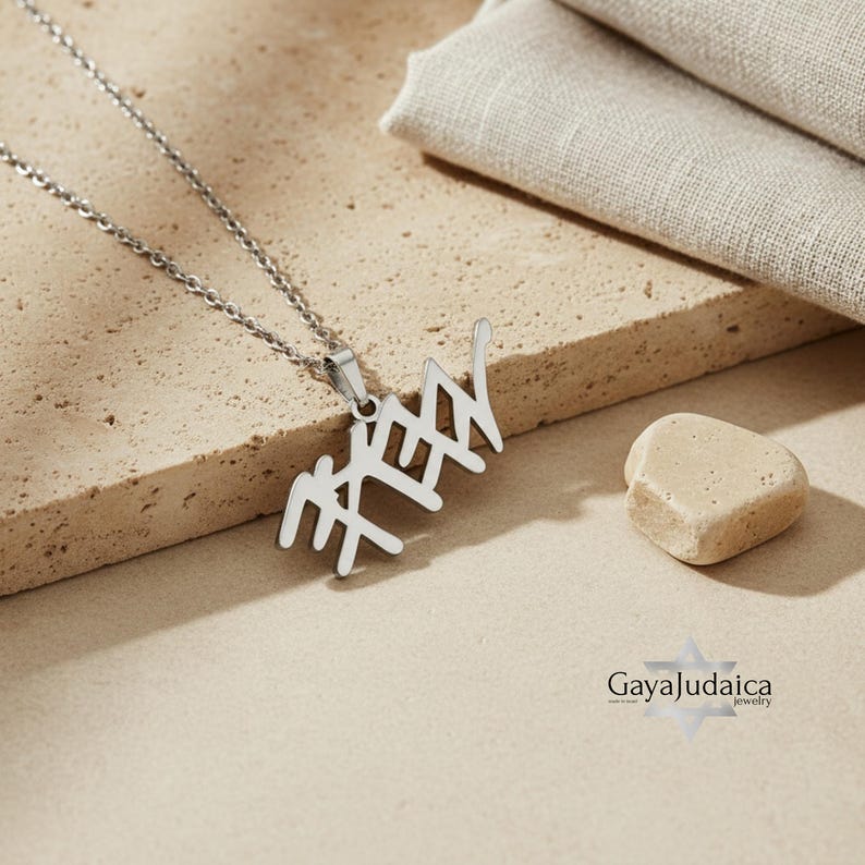 May include: A silver-colored pendant necklace featuring Hebrew script. The pendant hangs from a delicate chain and is displayed on a beige surface. A small, light-colored stone and a folded fabric are also in the image.