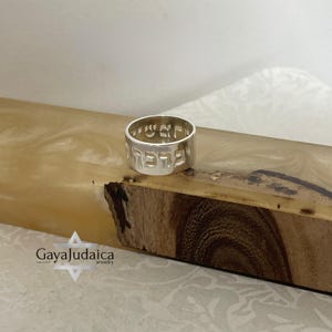 May include: A silver ring with Hebrew lettering cut out around the band. The ring is displayed on a wooden surface with a light-colored, marbled appearance. The ring is a piece of jewelry.