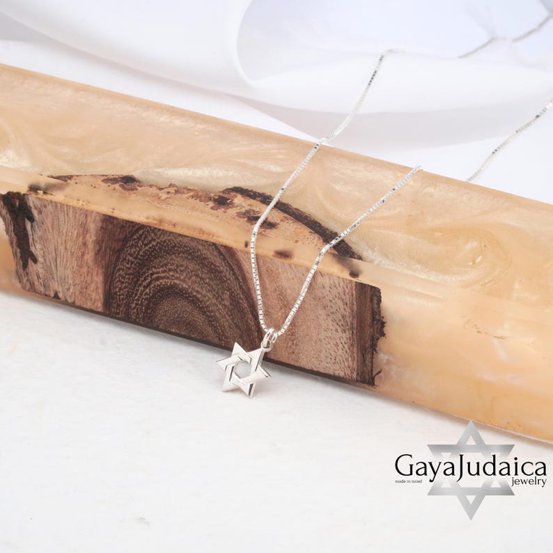 Pu&ograve; includere: Una collana a catena d'argento con un piccolo ciondolo a forma di Stella di David in argento. La collana &egrave; esposta su una superficie di legno. Il testo "Gaya Judaica Jewelry" &egrave; visibile nell'angolo in basso a destra dell'immagine.