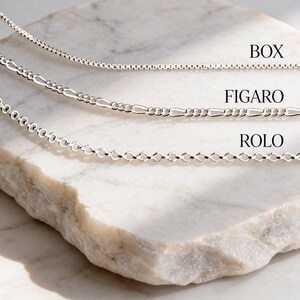 925 sterling silver box, figaro and rolo necklace chains draped over a white marble slab in soft natural light