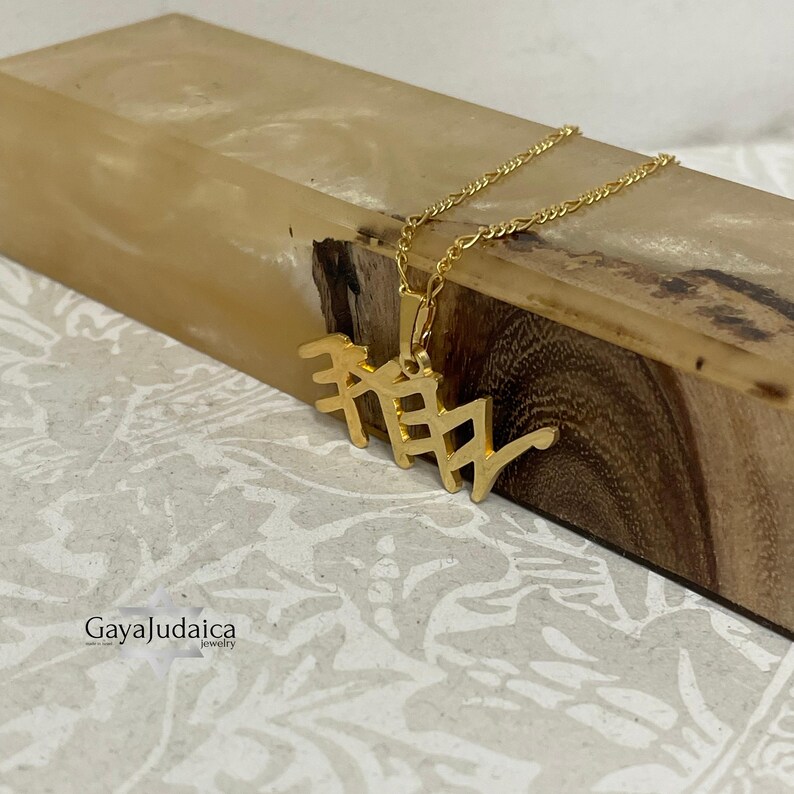 May include: A gold-colored pendant necklace with a Hebrew inscription. The pendant is suspended from a delicate gold chain. The necklace is displayed on a wooden block with a light-colored background. The brand name "GayaJudaica" is visible.