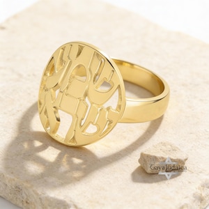 May include: A gold-colored ring with a circular design featuring cut-out Hebrew letters. The ring has a polished finish and sits on a light-colored stone surface. The ring's band is smooth and simple.