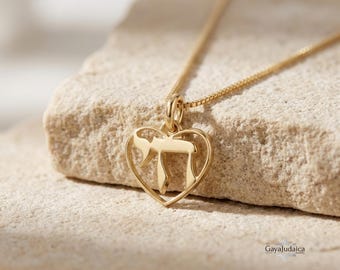 Puede incluir: Un collar con colgante dorado con un marco en forma de corazón y la palabra hebrea "Chai" en su interior. El collar se muestra sobre una superficie de piedra clara. La cadena es delicada y combina con el color del colgante. El diseño es elegante y simbólico.