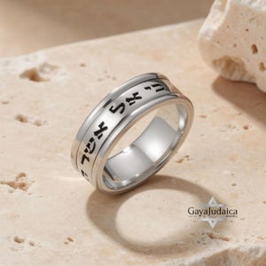 May include: A silver ring with Hebrew text engraved around the band. The ring has a polished finish and a simple, elegant design. The ring is displayed on a neutral-toned surface.