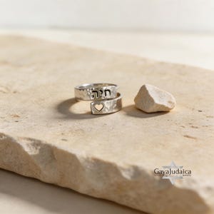 May include: Two silver rings with a hammered texture, one wrapped around the other. The top ring has Hebrew text, and the bottom ring features a heart cutout. A small, light-colored stone rests nearby.