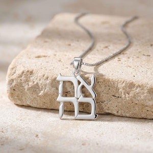 Puede incluir: Un collar con colgante de color plateado que presenta un dije rectangular con dos caracteres hebreos grabados en una fuente de estilo bloque. El colgante está suspendido de una cadena fina y delicada de color plateado con eslabones cuadrados.