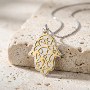 May include: A two-tone Hamsa hand pendant necklace featuring a gold-colored filigree design set against a silver-colored base. The pendant is attached to a delicate silver-colored box chain, showcasing intricate swirling patterns within the hand shape.