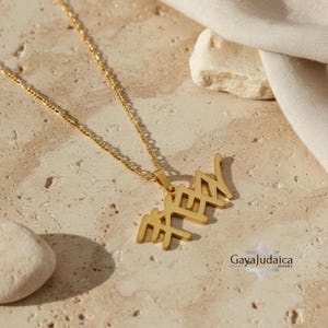 May include: A gold-colored necklace with a pendant featuring Hebrew letters. The necklace is displayed on a textured, light-brown surface. The chain is delicate, and the pendant is in a stylized font. The jewelry is from Gaya Judaica.