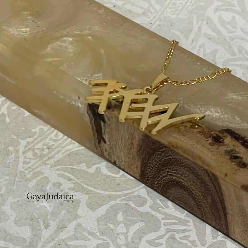 May include: A gold-colored pendant necklace featuring Hebrew script. The pendant is suspended from a delicate chain. The necklace is displayed on a wooden surface with a light-colored resin background. The Gaya Judaica logo is visible in the lower left corner.