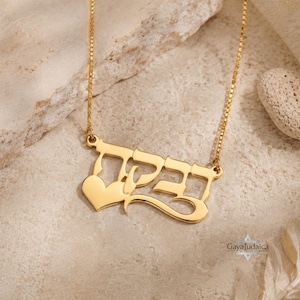 Puede incluir: Un collar dorado con un colgante con escritura hebrea y un corazón. La cadena es fina y el colgante es el punto focal sobre un fondo neutro. El diseño es elegante y el estilo general es minimalista.