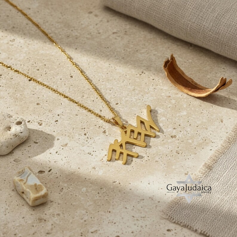 May include: A gold-colored necklace with a pendant featuring Hebrew letters. The necklace is displayed on a textured, light-colored surface, with small stones and a dried leaf nearby. The chain is delicate and the pendant is the focal point.