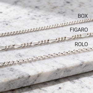 Silver box, figaro and rolo chains on gray marble, three classic 925 sterling silver necklace chain styles shown together