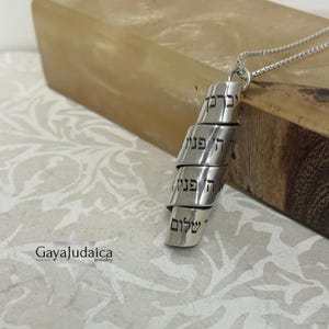 May include: A silver-colored pendant necklace with Hebrew text. The pendant is a spiraling design with the words etched into the metal. The necklace is displayed on a wooden surface. The logo "Gaya Judaica jewelry" is visible in the lower left corner.