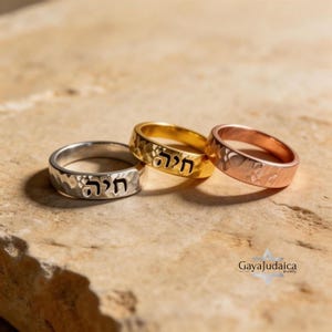May include: Three hammered-finish rings in silver, gold, and rose gold. Each ring features Hebrew text. The rings are displayed on a textured, light brown surface. The rings are from Gaya Judaica jewelry.