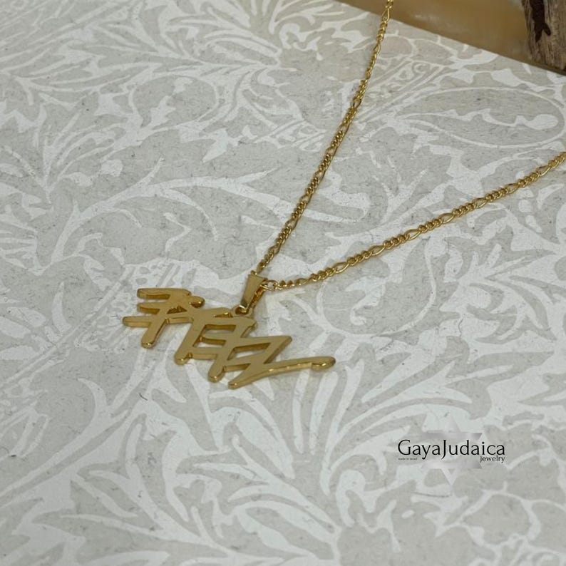 May include: Gold-colored pendant necklace with Hebrew text. The pendant is in a stylized font and hangs from a delicate chain. The necklace is displayed on a patterned surface. The brand name "GayaJudaica" is visible.