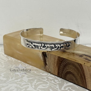 May include: A silver cuff bracelet with Hebrew text. The bracelet is displayed on a wooden block with a light-colored background. The text is in a stylized font and is centered on the bracelet. The bracelet is open at the ends.
