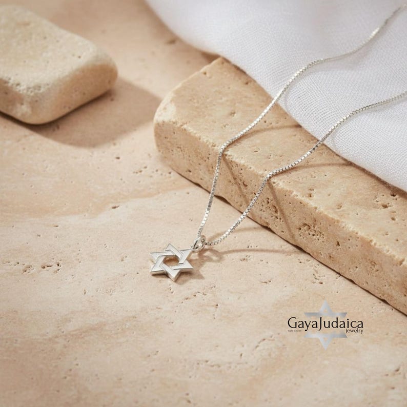 May include: A silver chain necklace with a small silver Star of David pendant. The necklace is displayed on a wooden surface. The text "Gaya Judaica Jewelry" is visible in the bottom right corner of the image.