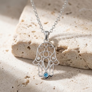May include: A silver-toned Hamsa hand pendant necklace featuring intricate openwork filigree detailing and a single small, round, bright blue gemstone set at the center of the palm. The pendant hangs from a delicate silver-toned link chain.