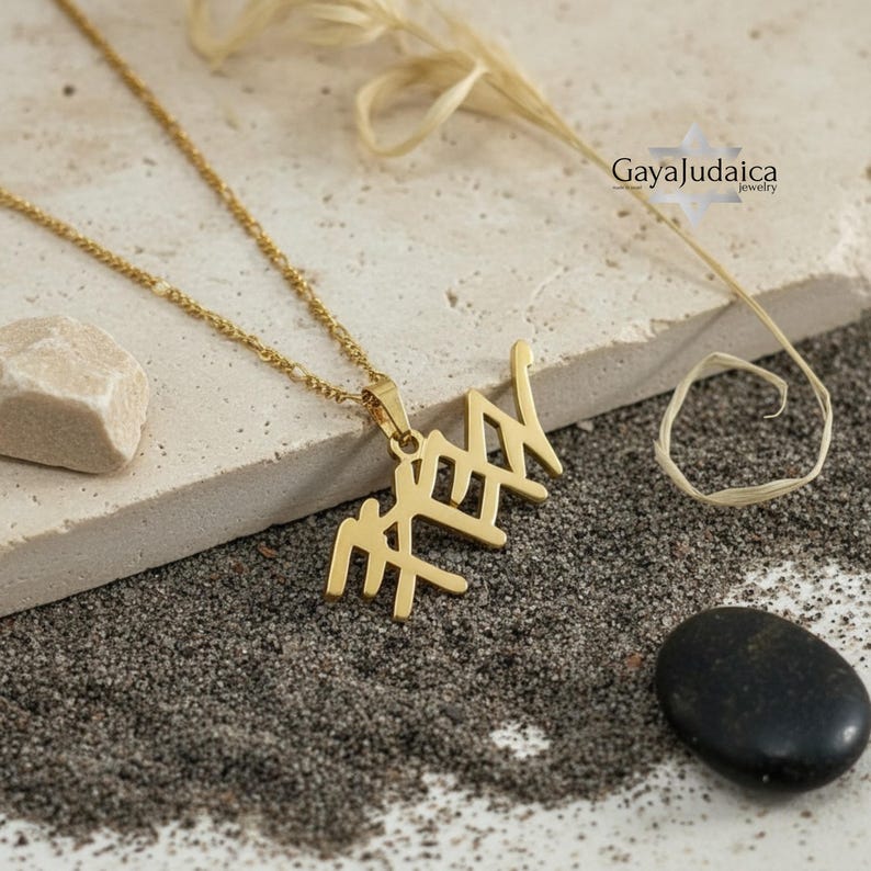May include: A gold-colored pendant necklace with a Hebrew inscription. The pendant hangs from a delicate chain. The necklace is displayed on a textured surface with a small rock and a black stone. The brand name "GayaJudaica" is visible in the upper right corner.