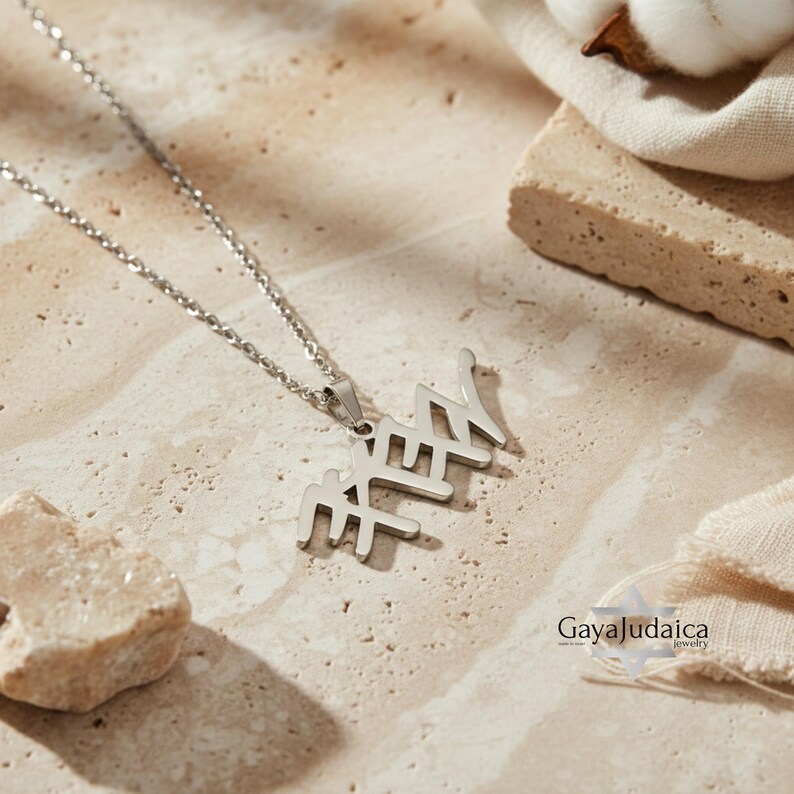 May include: A silver-colored pendant necklace with Hebrew letters. The necklace is displayed on a beige surface with a cotton ball and a piece of fabric in the background. The necklace is from Gaya Judaica jewelry.