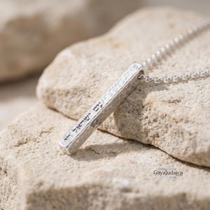 May include: A silver bar pendant necklace with Hebrew text. The pendant has a hammered texture and hangs from a delicate chain. The necklace is displayed on a light-colored stone surface. The jewelry is from Gaya Judaica.