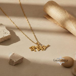 May include: A gold-colored necklace with a pendant featuring Hebrew script. The pendant hangs from a delicate chain. The necklace is displayed on a neutral-toned surface with decorative stones and a rolled paper element. The logo "Gaya Judaica" is visible.