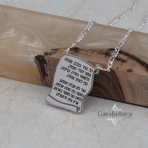 Puede incluir: Collar con colgante plateado con diseño de pergamino con texto en hebreo. El colgante cuelga de una delicada cadena. El colgante se muestra sobre un fondo de madera, con el nombre de la marca "GayaJudaica".