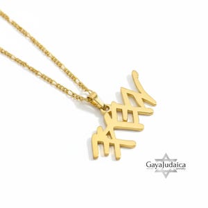 May include: A gold-colored pendant necklace with Hebrew script spelling out the word "Chai". The pendant is attached to a delicate gold chain. The necklace is displayed on a white background.